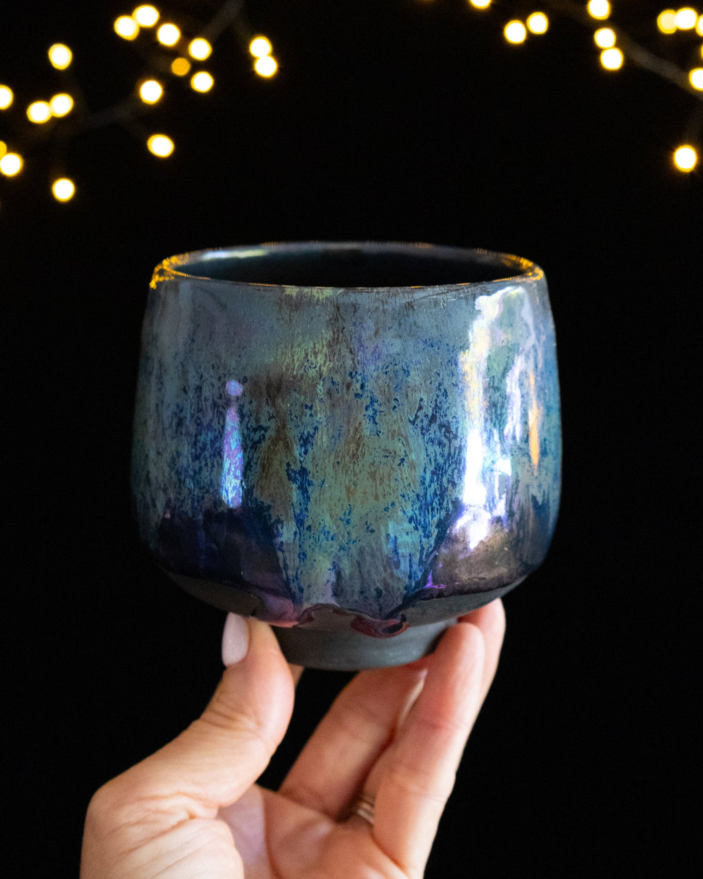 Handmade lusterware ritual cup in soft teal and violet, held against fairy lights