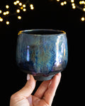 Handmade lusterware ritual cup in soft teal and violet, held against fairy lights