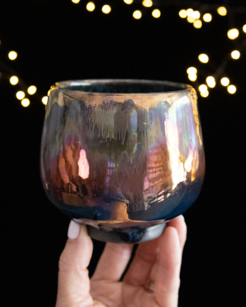 Handmade lusterware ritual cup in warm rose and bronze, held against fairy lights