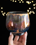Handmade lusterware ritual cup in warm rose and bronze, held against fairy lights